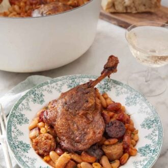 Pinterest graphic of a plate with a serving of cassoulet with a pot with the rest in the background with some wine and bread.