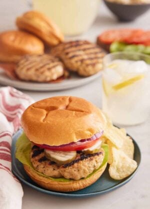 A plate with a chicken burger with potato chips with a drink in the background and burger components on plates.