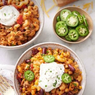 Pinterest graphic of bowls of chili mac topped with sour cream with a bowl of sliced jalapeño and sliced limes.