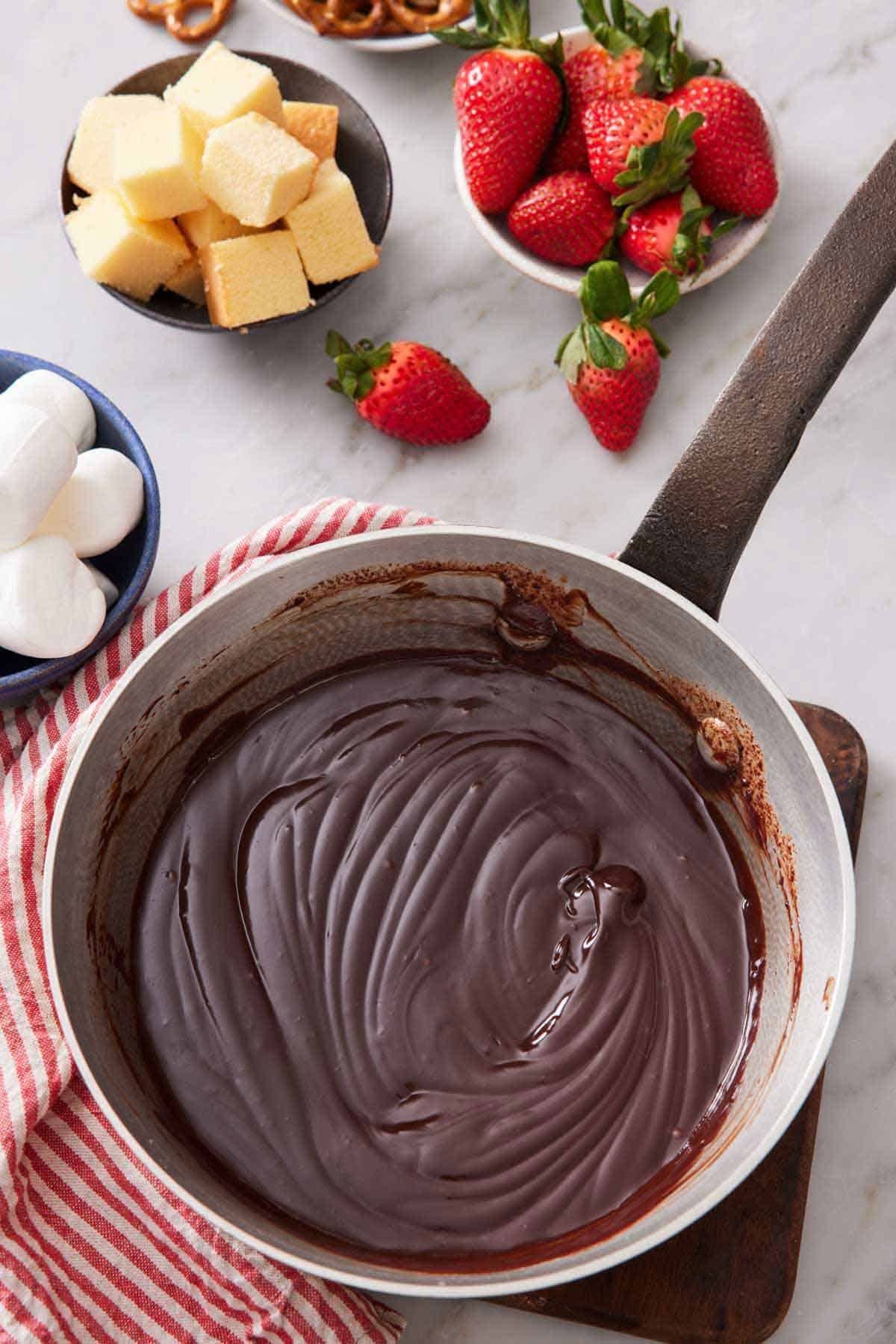 A saucepan of chocolate fondue with bowls of multiple dipper options in the background.