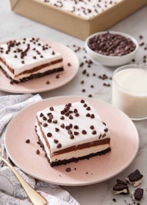 Two plates with sliced chocolate lasagna with a glass of milk, bowl of mini chocolate chips, and a pan of more chocolate lasagna in the background.