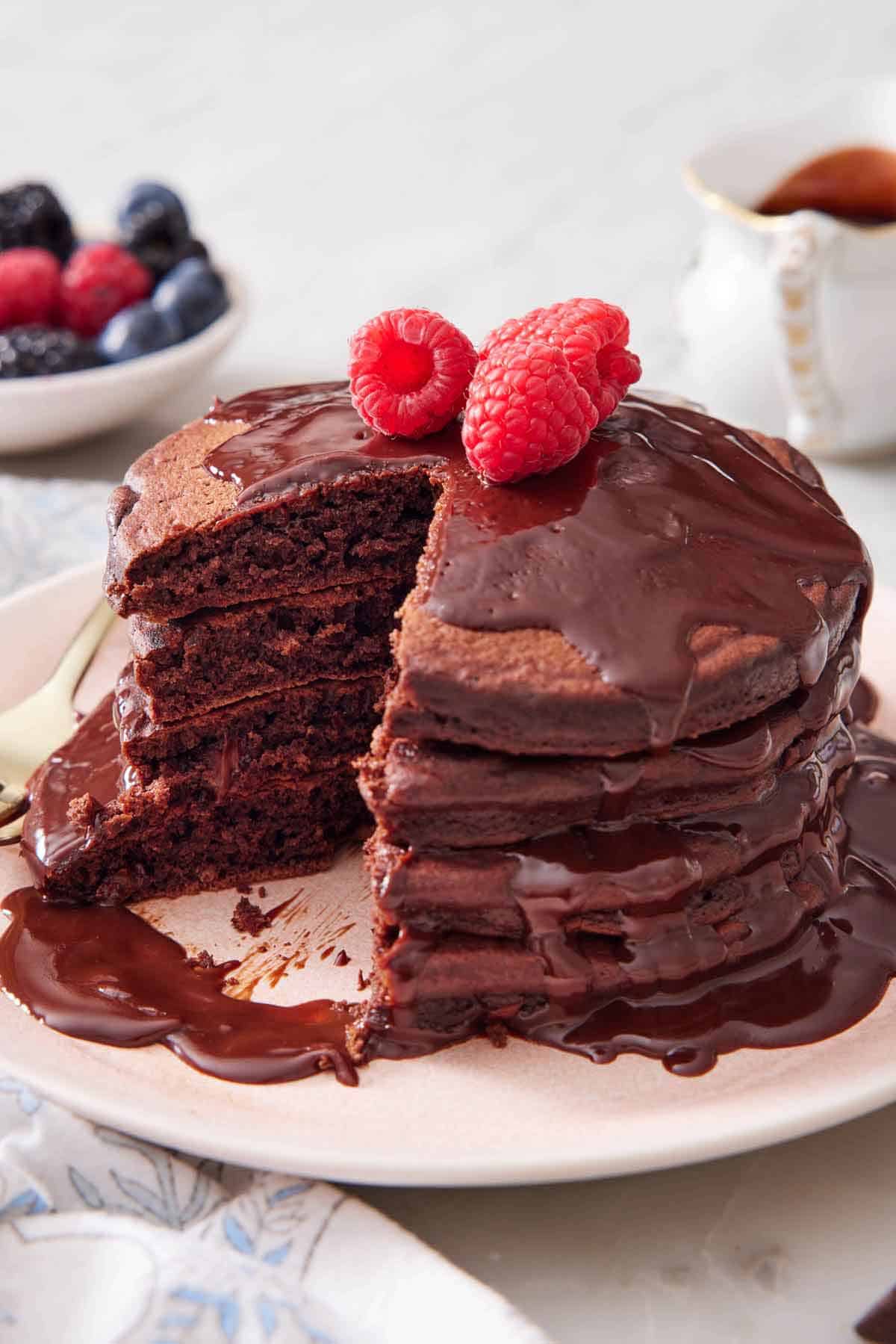 A stack of chocolate pancakes topped with chocolate sauce and raspberries with a fifth cut out.