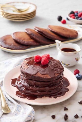 A plate with a stack of chocolate pancakes with chocolate sauce and topped with raspberries. More pancakes and sauce in the background.