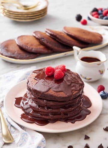 A plate with a stack of chocolate pancakes with chocolate sauce and topped with raspberries. More pancakes and sauce in the background.