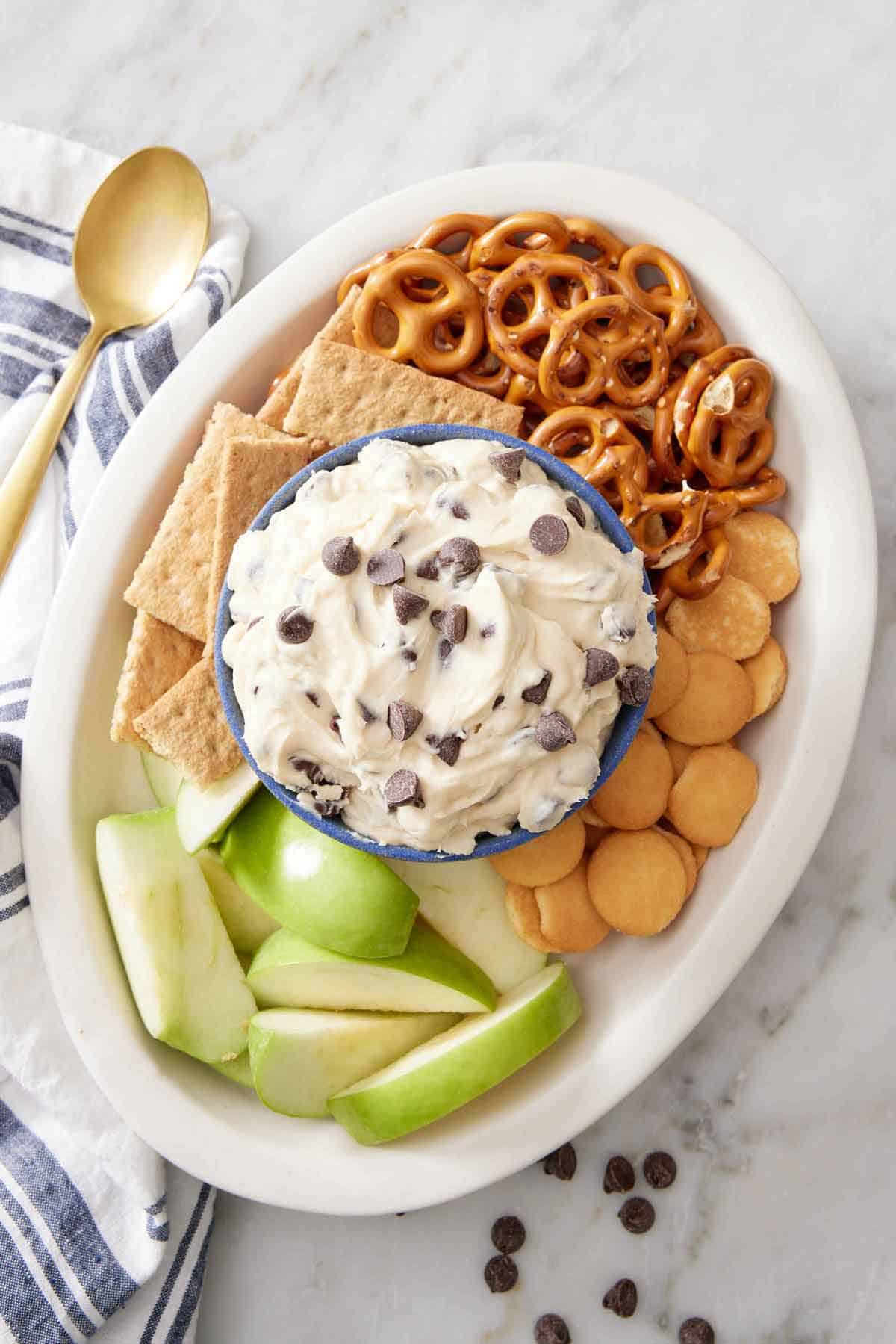 Overhead view of a platter with a bowl of cookie dough dip surrounded by crackers, pretzels, and sliced apples.