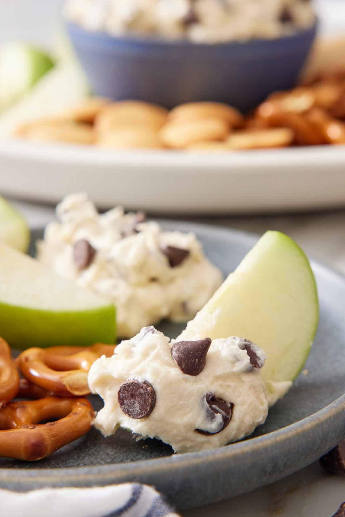 A plate with slice of apple dipped in cookie dough dip. More dip and dippers in the background.