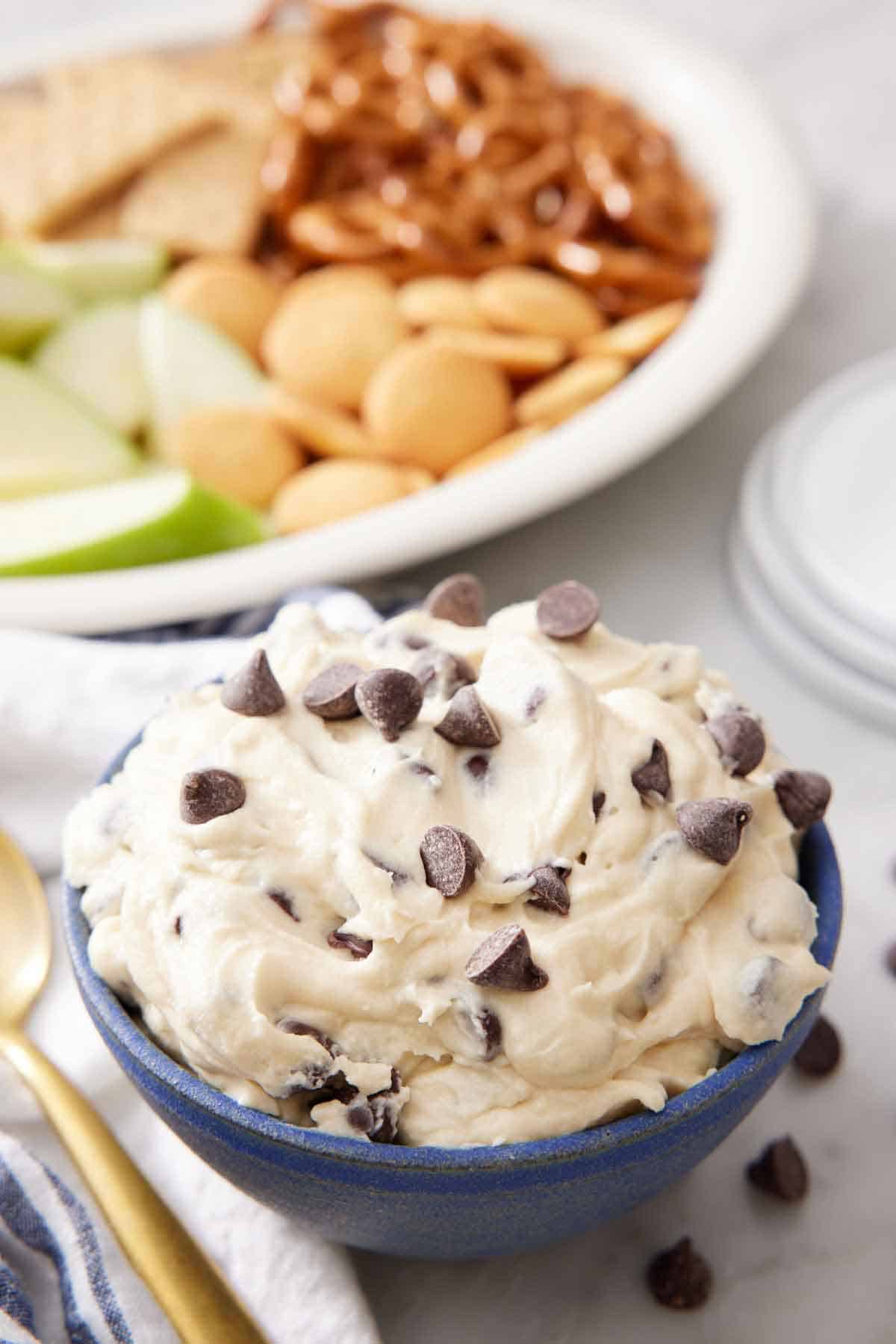 A bowl of cookie dough dip with crackers, pretzels, and apples in the background.