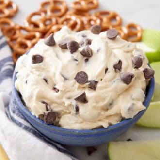 A bowl of cookie dough dip surrounded by apple slices and pretzels with a napkin and spoon.