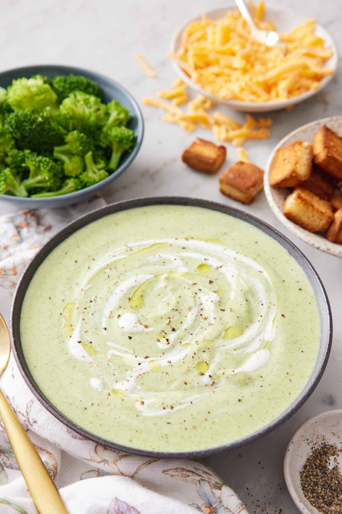 A bowl of cream of broccoli soup with cream swirled on top with pepper. Broccoli, shredded cheese, and croutons in the background.