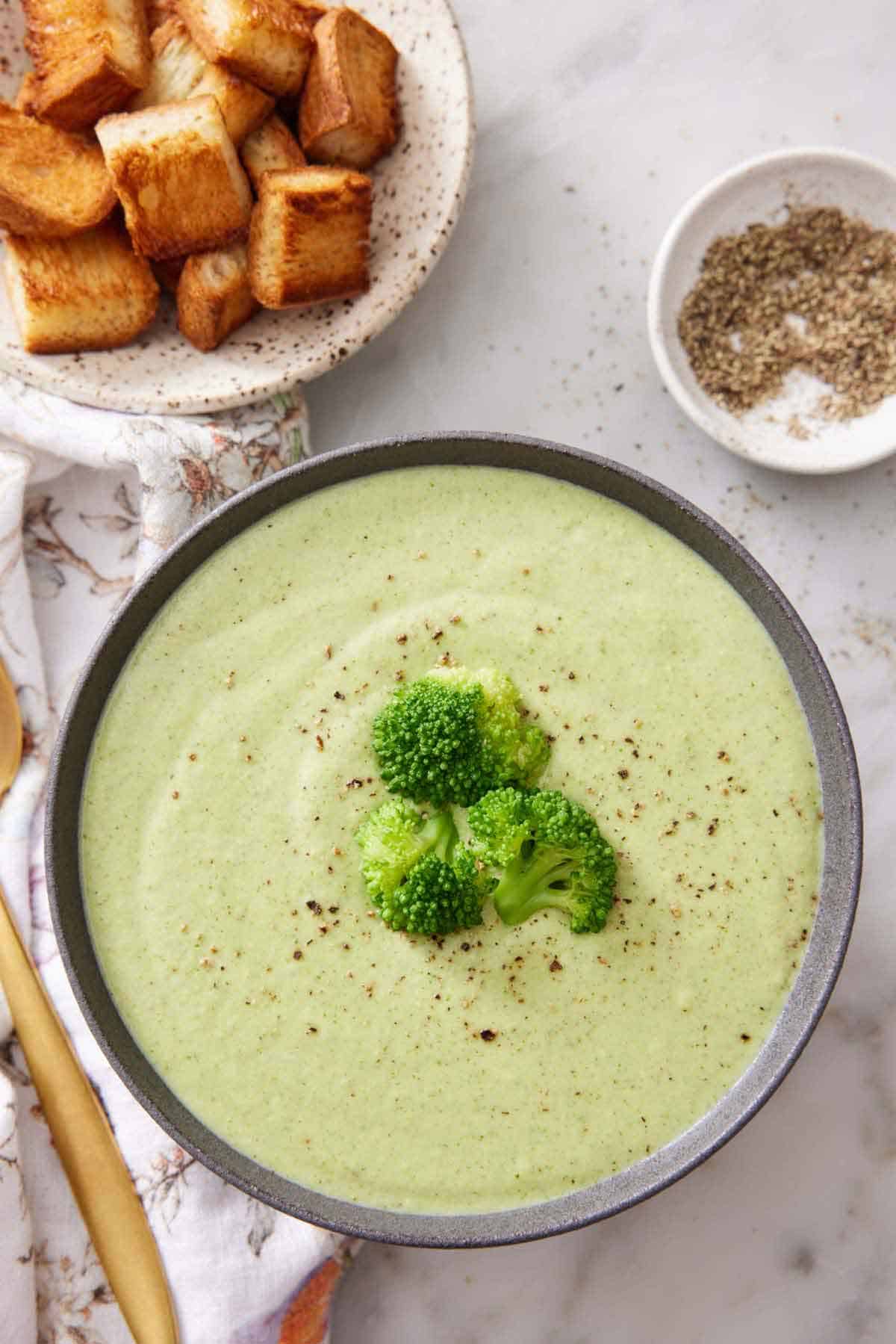 Overhead view of a bowl of cream of broccoli soup topped with three florets and some black pepper. Bowl of pepper and croutons off to the side.