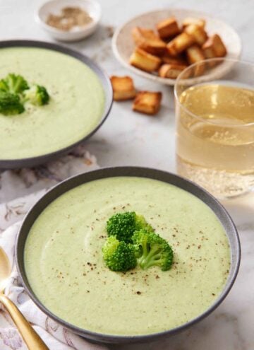 Two bowls of cream of broccoli soup with one front and center. Both bowls topped with fresh broccoli florets and pepper. A glass of wine in the back with croutons.