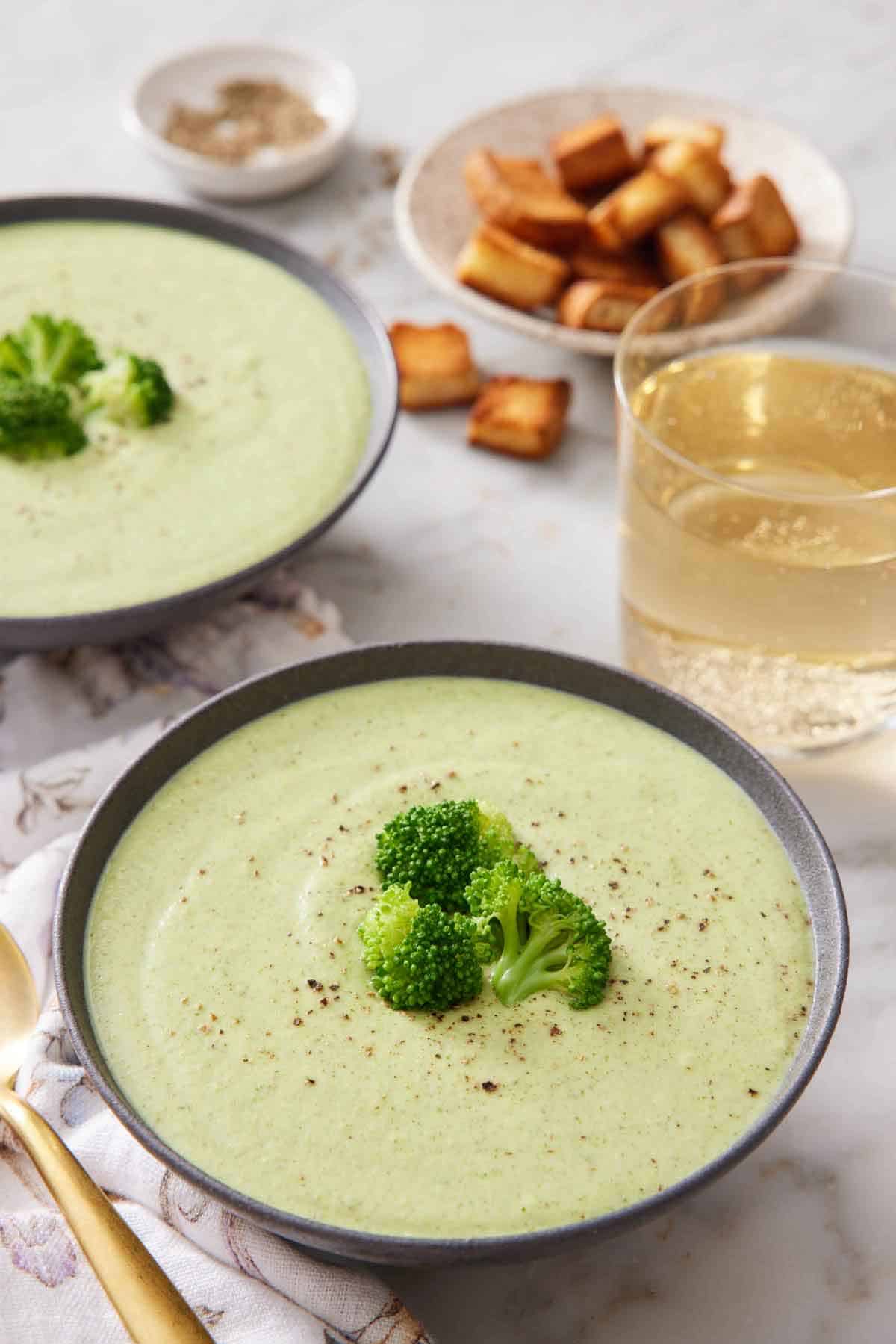 Two bowls of cream of broccoli soup with one front and center. Both bowls topped with fresh broccoli florets and pepper. A glass of wine in the back with croutons.