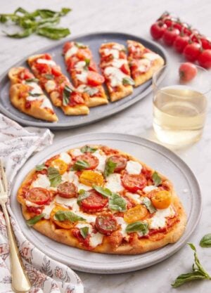 A plate with a flatbread pizza with a second plate in the background, cut into long slices. Glass of wine, tomatoes, and basil scattered around.