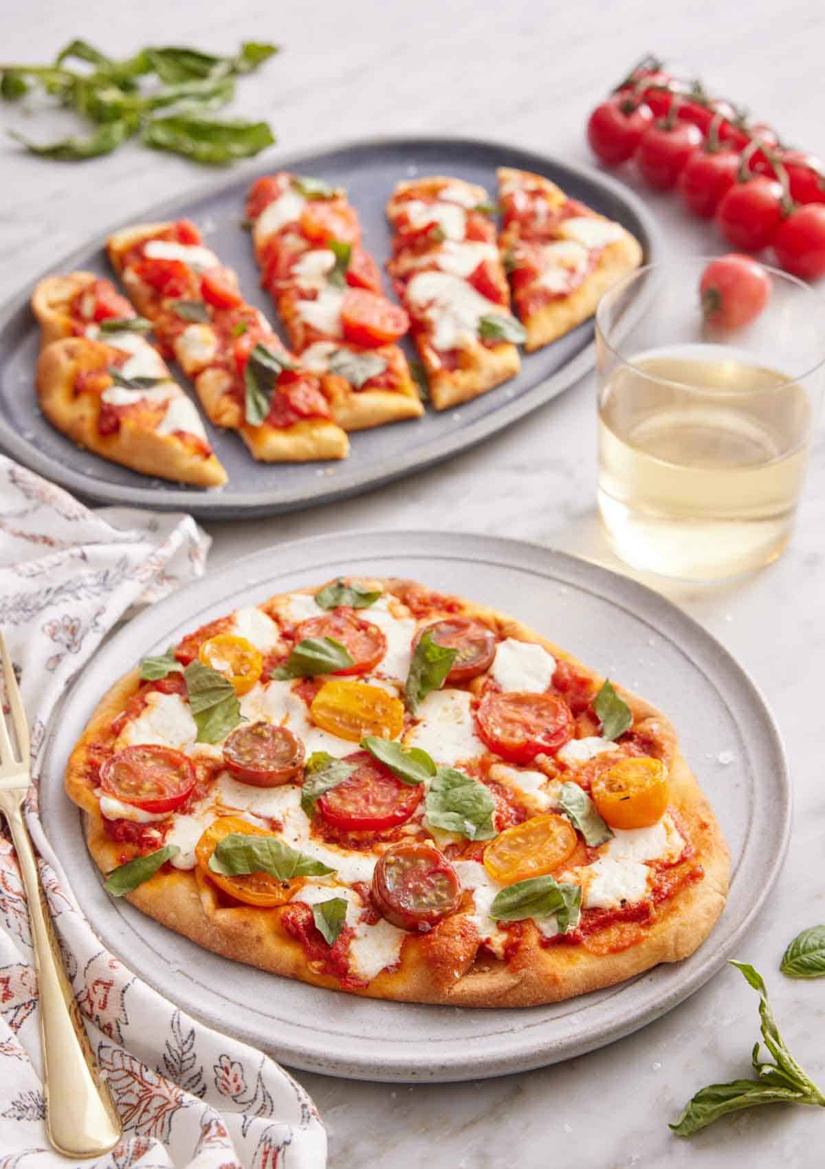A plate with a flatbread pizza with a second plate in the background, cut into long slices. Glass of wine, tomatoes, and basil scattered around.