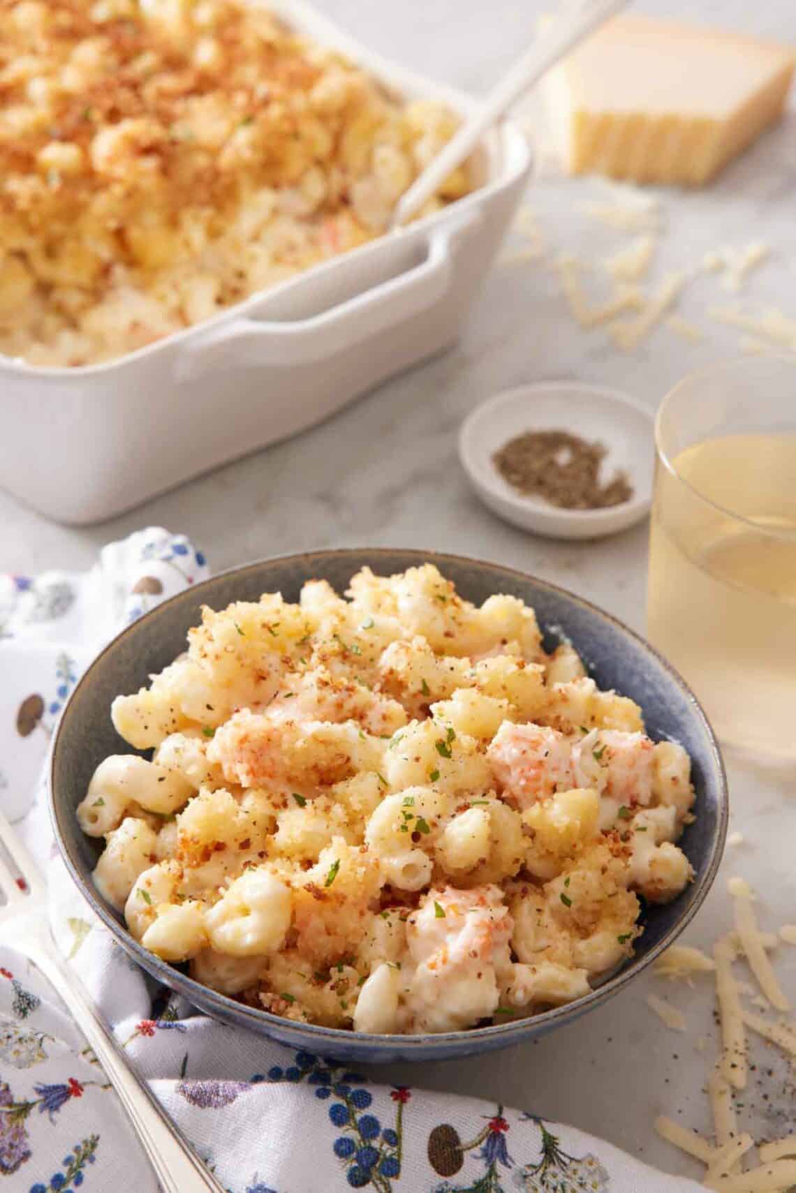A bowl of lobster mac and cheese with a baking dish with more in the background along with a drink and bowl of pepper.
