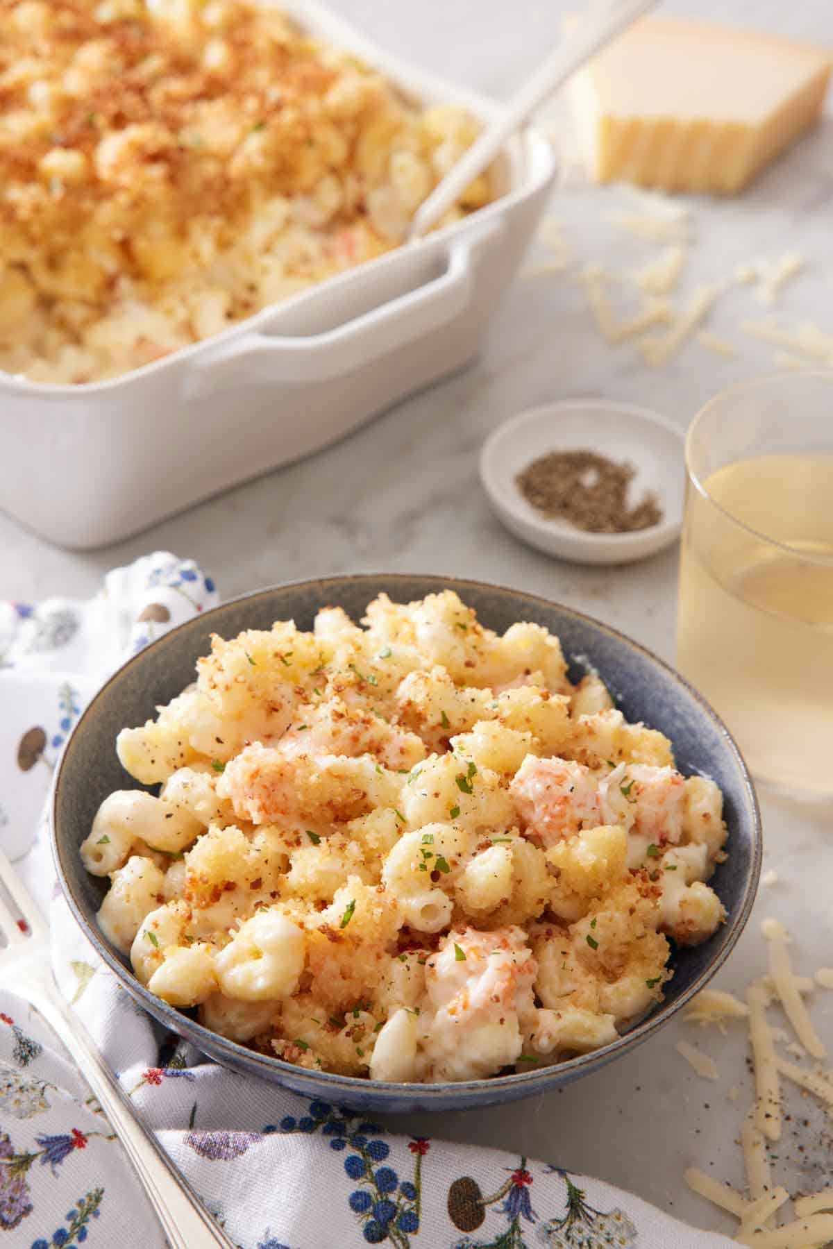 A bowl of lobster mac and cheese with a baking dish with more in the background along with a drink and bowl of pepper.