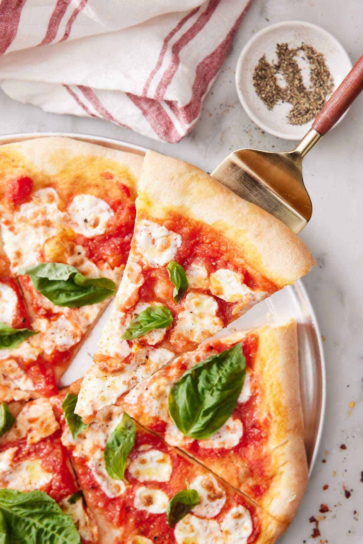 A slice of margherita pizza being lifted from the fresh of the pizza. A small bowl of pepper and a linen beside it.