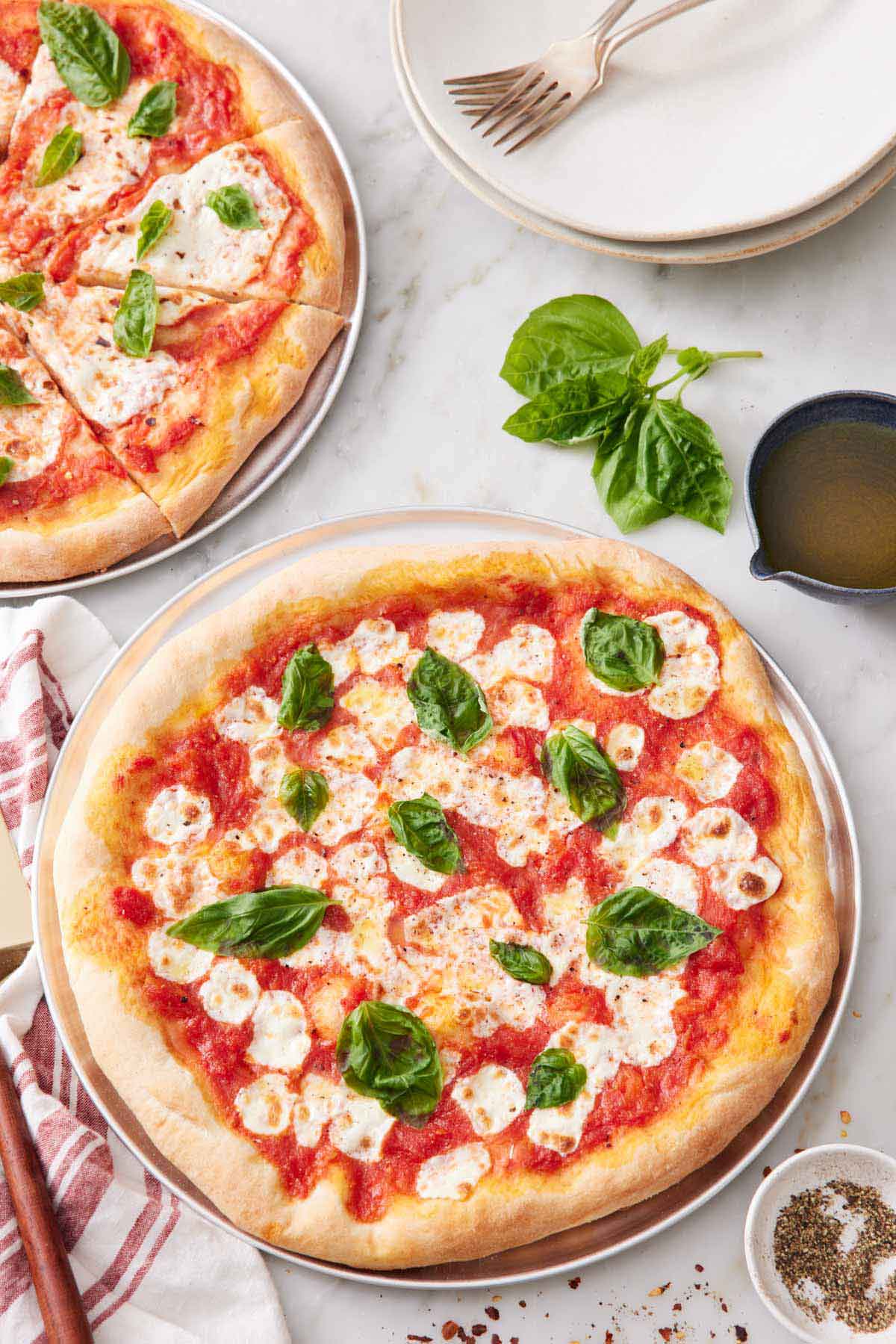 A margherita pizza topped with fresh basil leaves with a second sliced pizza in the background along with a stack of plates and forks.