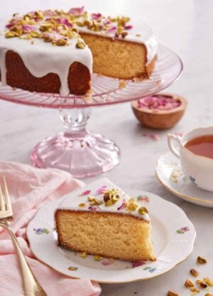 A slice of Persian love cake on a plate with a pink cake stand in the back with the rest of the cake.