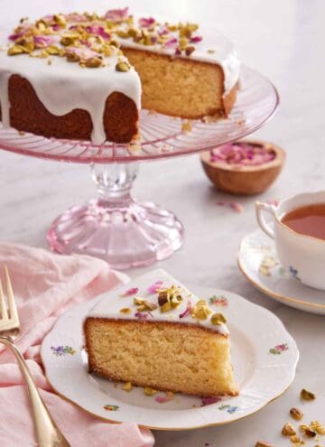 A slice of Persian love cake on a plate with a pink cake stand in the back with the rest of the cake.