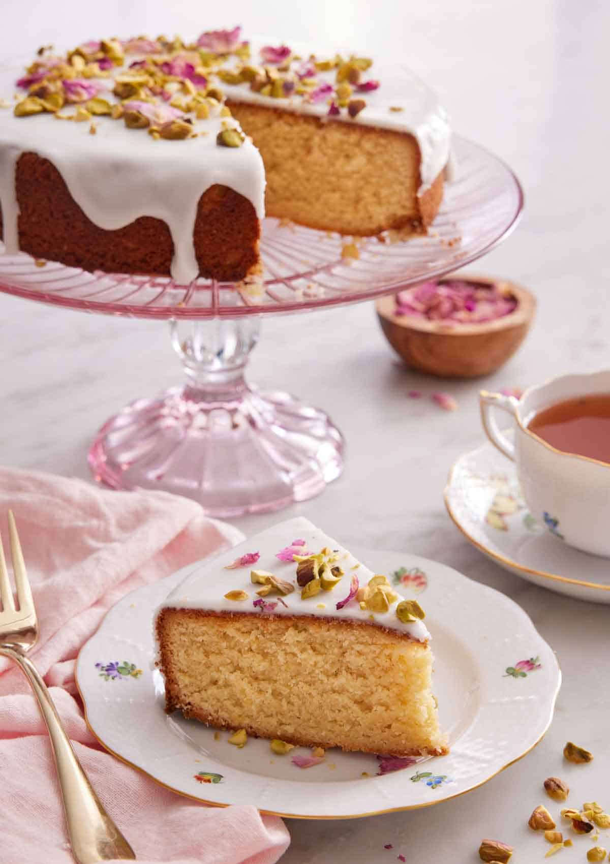 A slice of Persian love cake on a plate with a pink cake stand in the back with the rest of the cake.