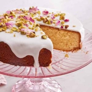 A pik cake stand with a Persian love cake with a slice removed. A small bowl of dried rose petals in the background.