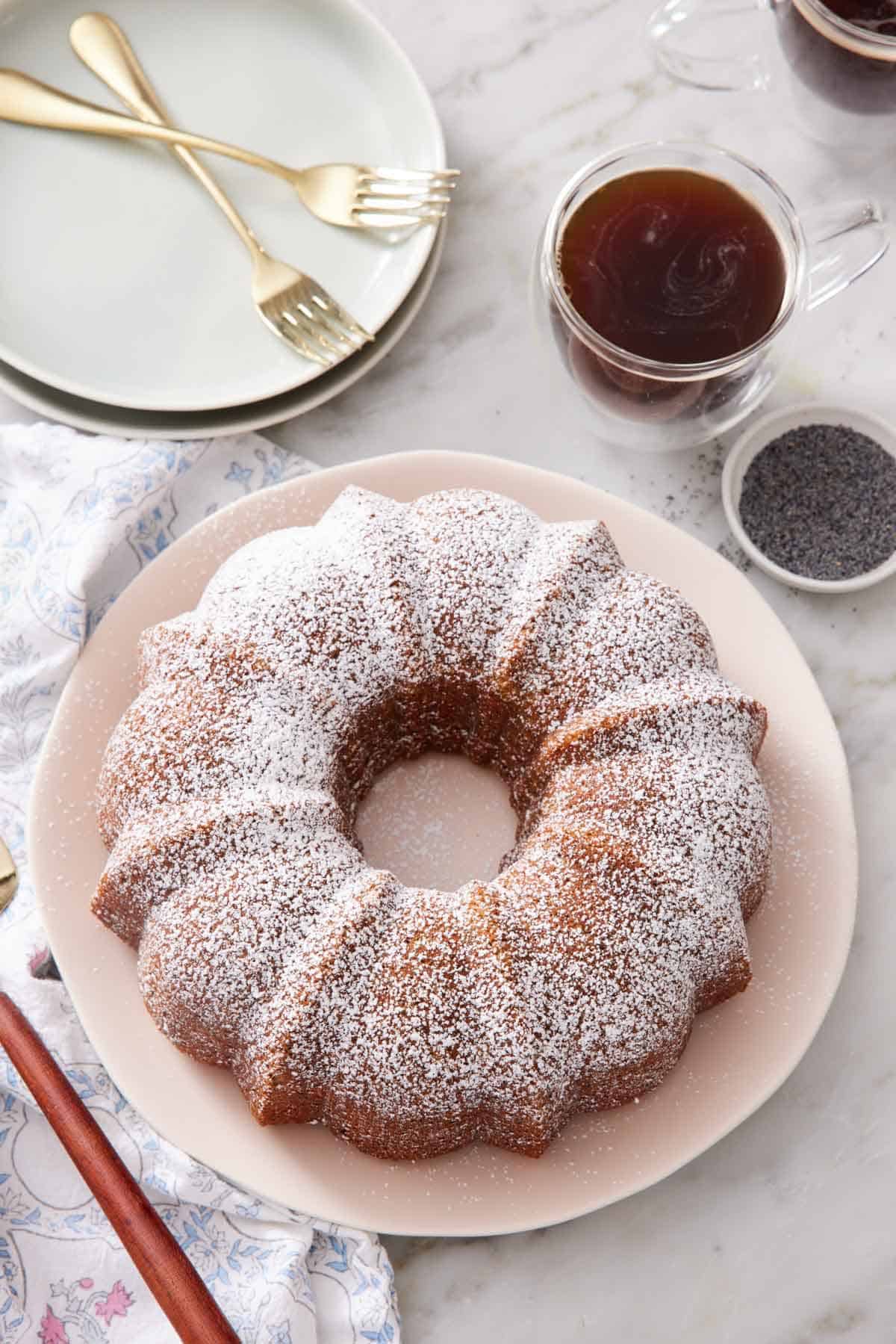 Overhead view of a poppy seed cake on a platter dusted with powdered sugar. Coffee and poppy seed in the back with some plates and forks.