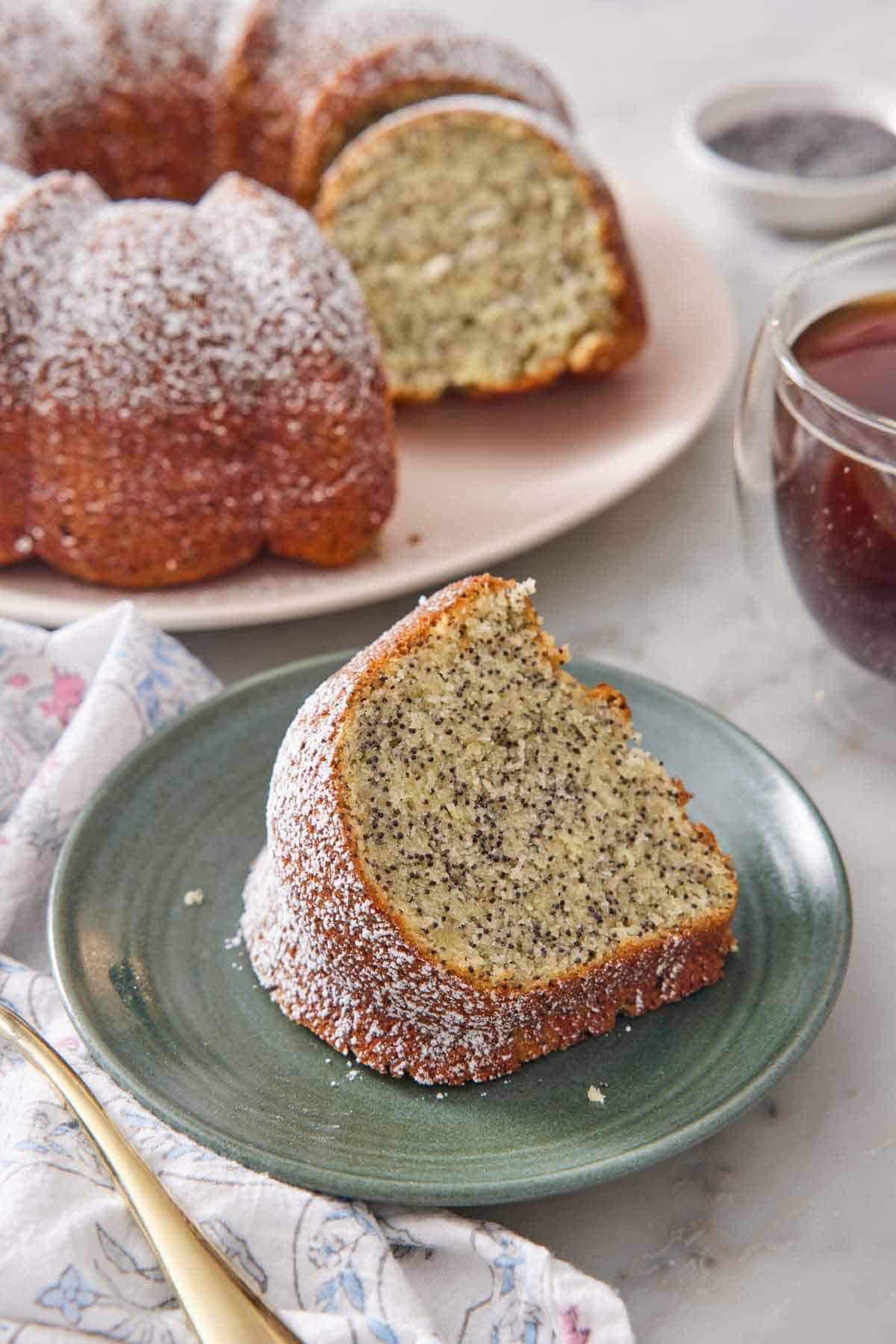 A slice of poppy seed cake on a green plate with the rest of the cake in the background.