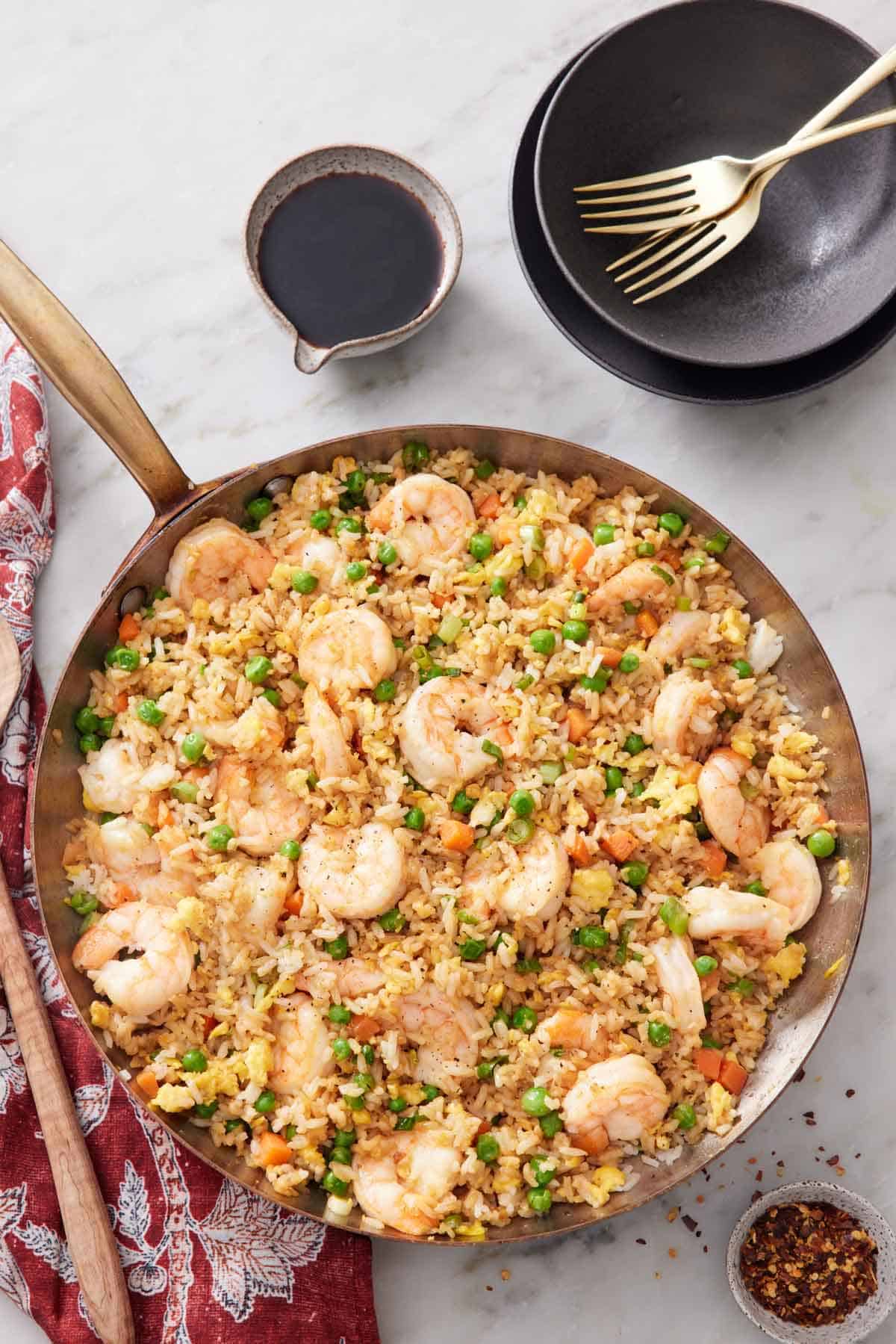 An overhead view of a skillet of shrimp fried rice with a stack of bowls, forks, some soy sauce, and some red pepper flakes off to the side.