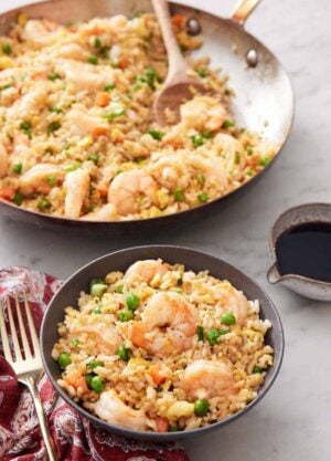 A bowl of shrimp fried rice with a skillet with more in the background with a wooden spoon.