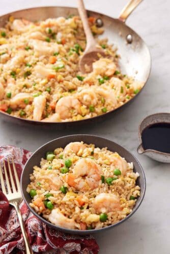 A bowl of shrimp fried rice with a skillet with more in the background with a wooden spoon.