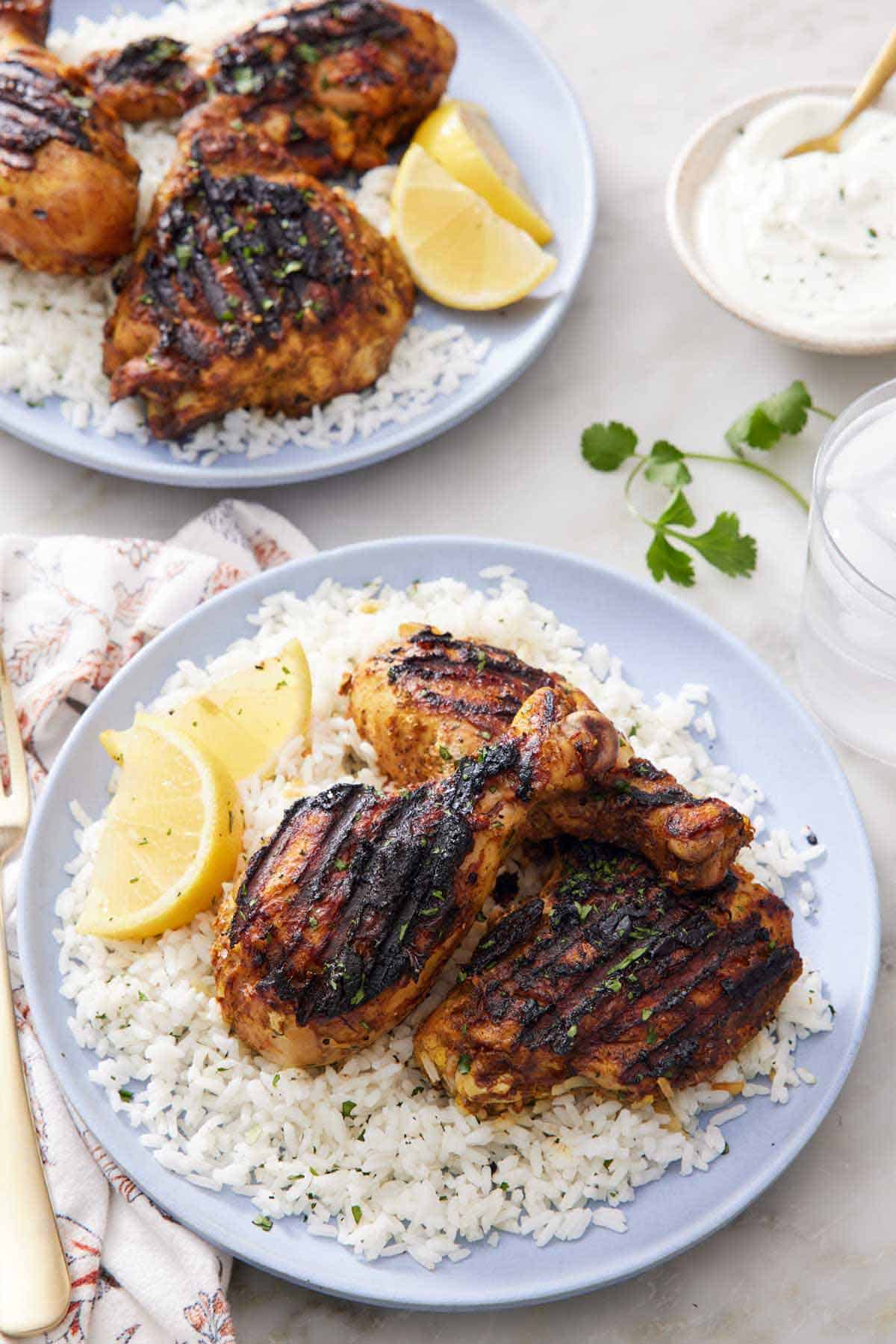 A plate with tandoori chicken over a bed of rice with lemon wedges. A second plate in the back along with a white sauce.