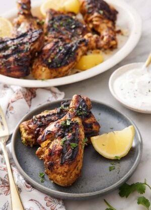 A plate with two pieces of tandoori chicken with a lemon wedge with a yogurt sauce in the background with the rest of the chicken on a platter.