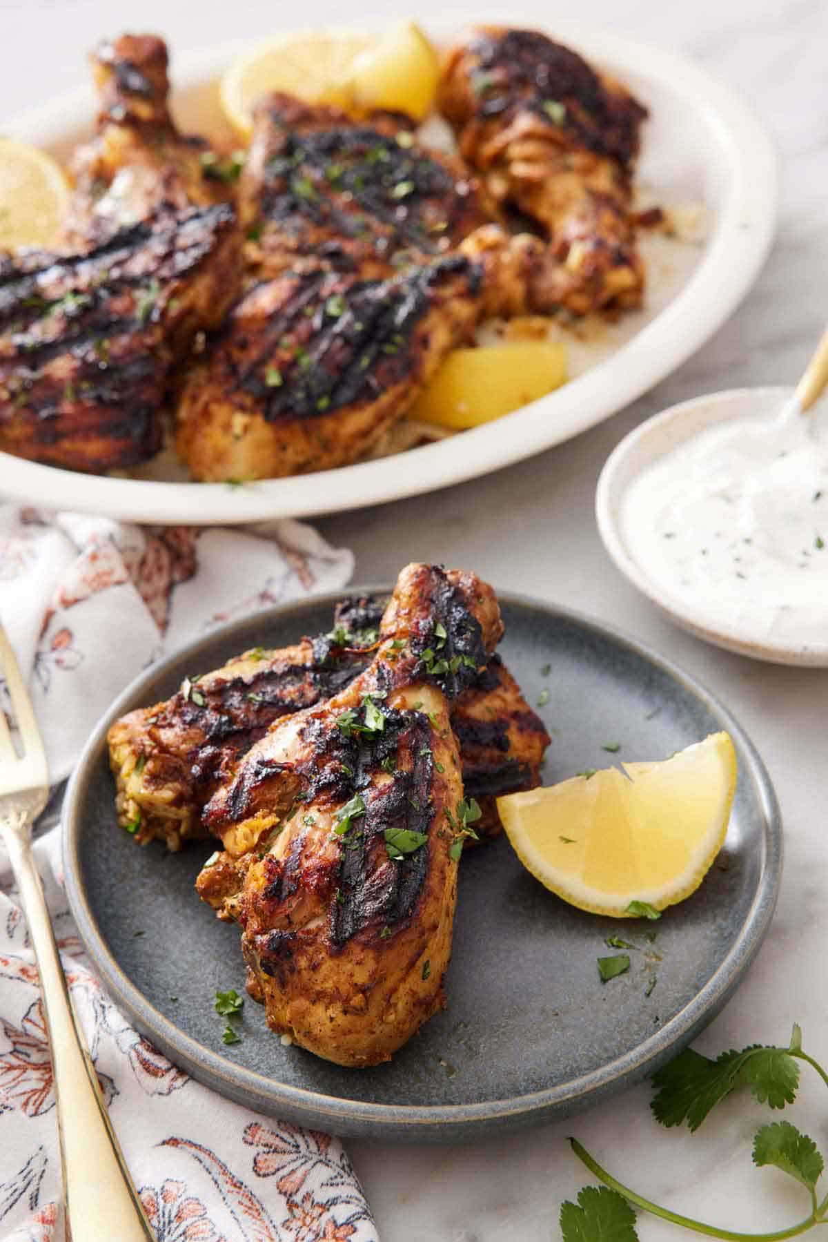 A plate with two pieces of tandoori chicken with a lemon wedge with a yogurt sauce in the background with the rest of the chicken on a platter.