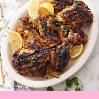 Pinterest graphic of a platter of tandoori chicken topped with chopped parsley with lemon wedges. Yogurt sauce in the back with a stack of plates.