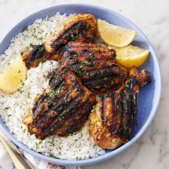 A plate of tandoori chicken over rice with chopped parsley and lemon wedges.