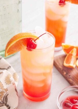A glass of tequila sunrise with an orange slice and maraschino cherry as garnish. A second glass in the background along with the cut orange slices and tequila bottle.