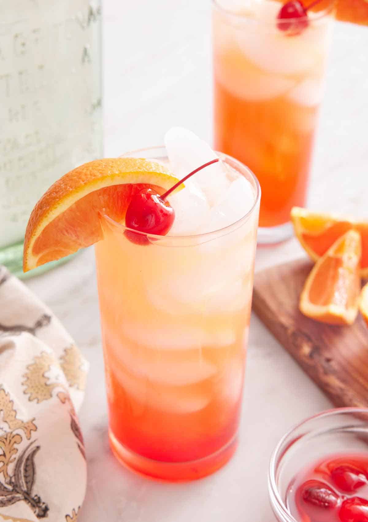 A glass of tequila sunrise with an orange slice and maraschino cherry as garnish. A second glass in the background along with the cut orange slices and tequila bottle.