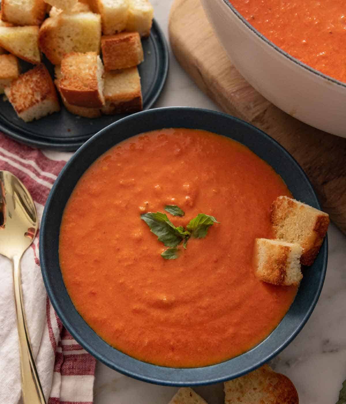 Overhead view of a bowl of tomato soup with basil and bread on top. A plate of bread off to the side along with a pot of the soup.