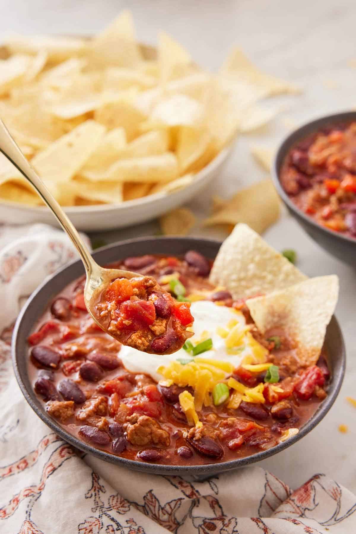 A spoonful of turkey chili lifted from a bowl. A plate of chips in the background.