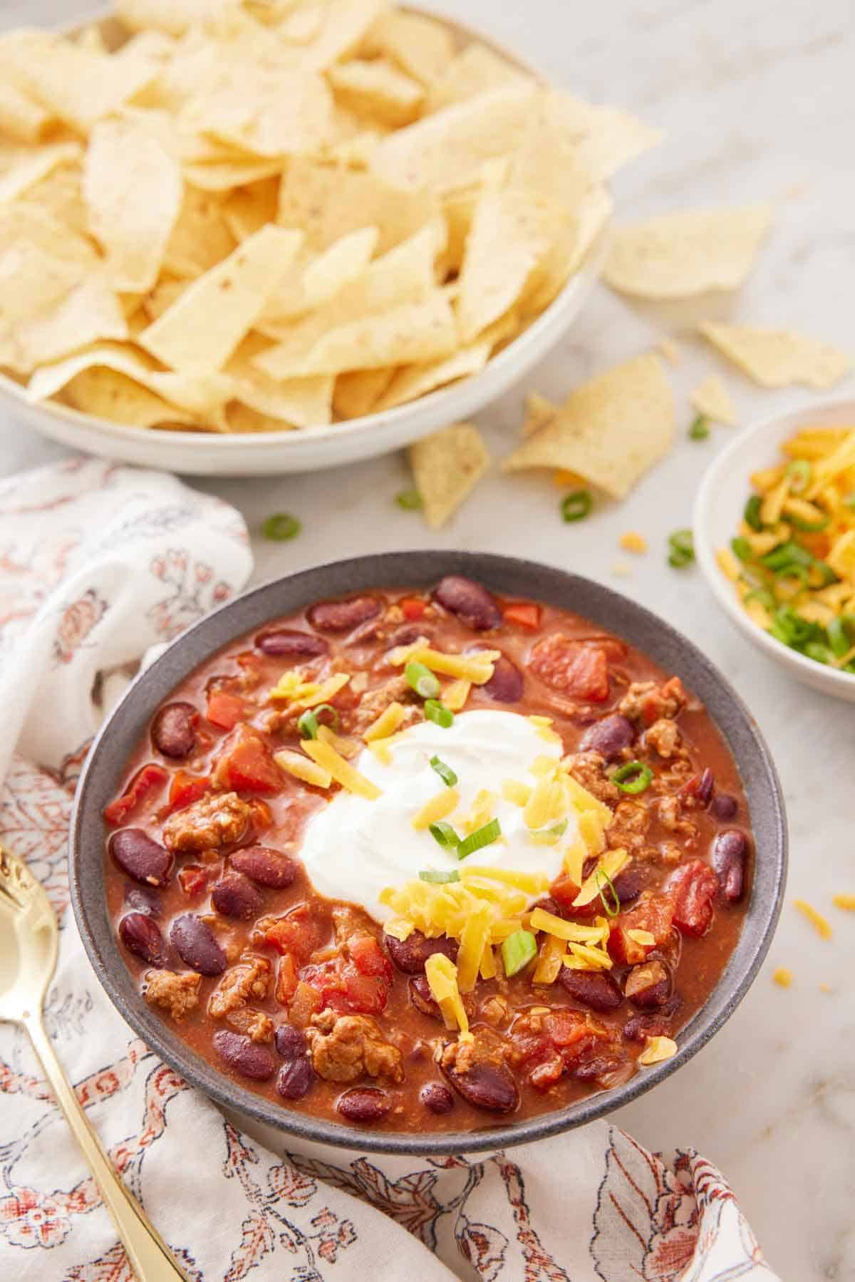 A bowl of turkey chili topped with sour cream and cheese with a plate of chips and bowl of toppings in the back.
