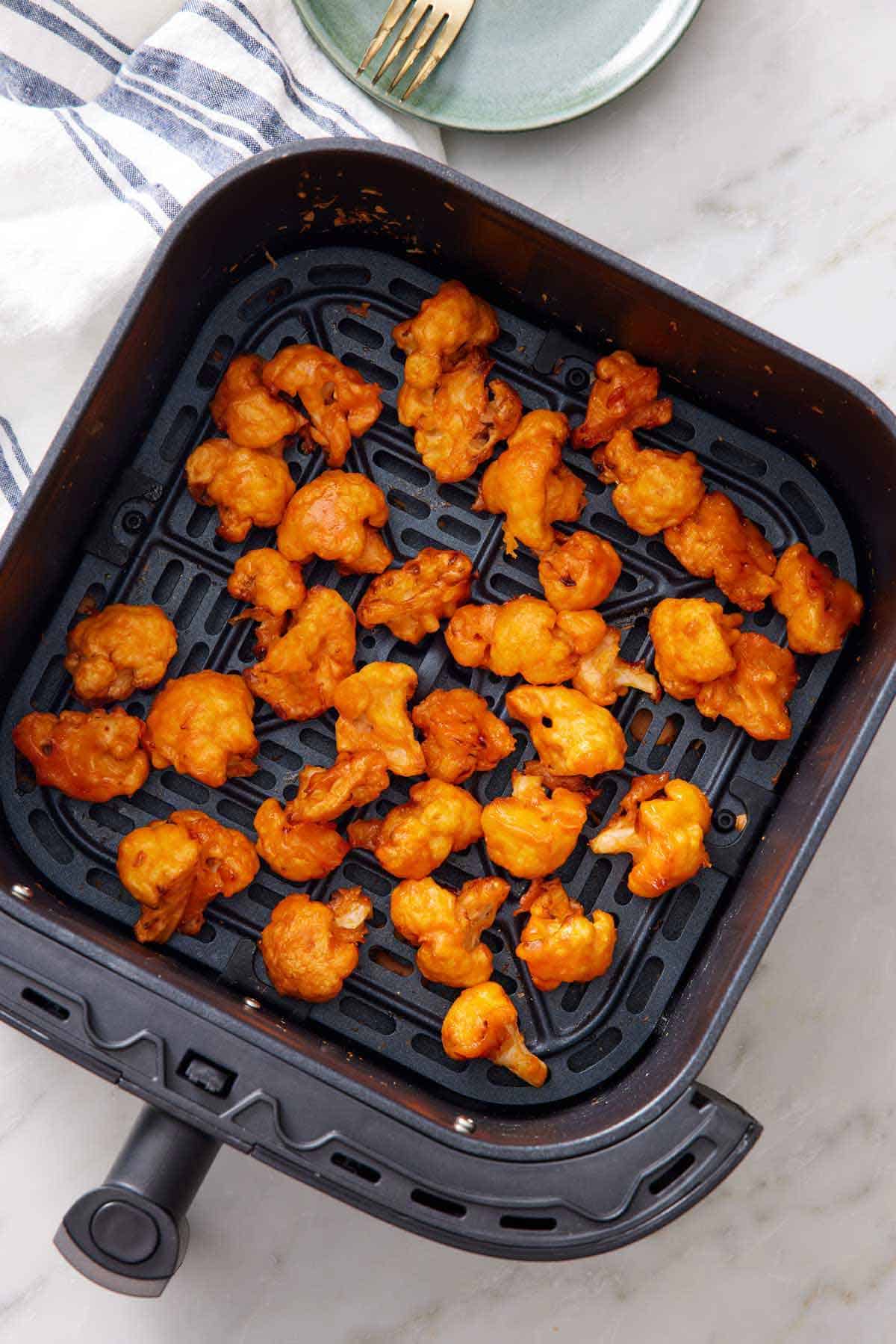 Overhead view of buffalo cauliflower in the air fryer basket. A napkin, plate, and fork on the side.