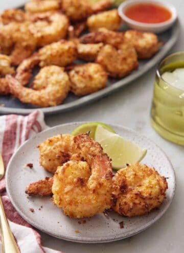 A plate of air fryer coconut shrimp with lime. A platter with more shrimp in the background.