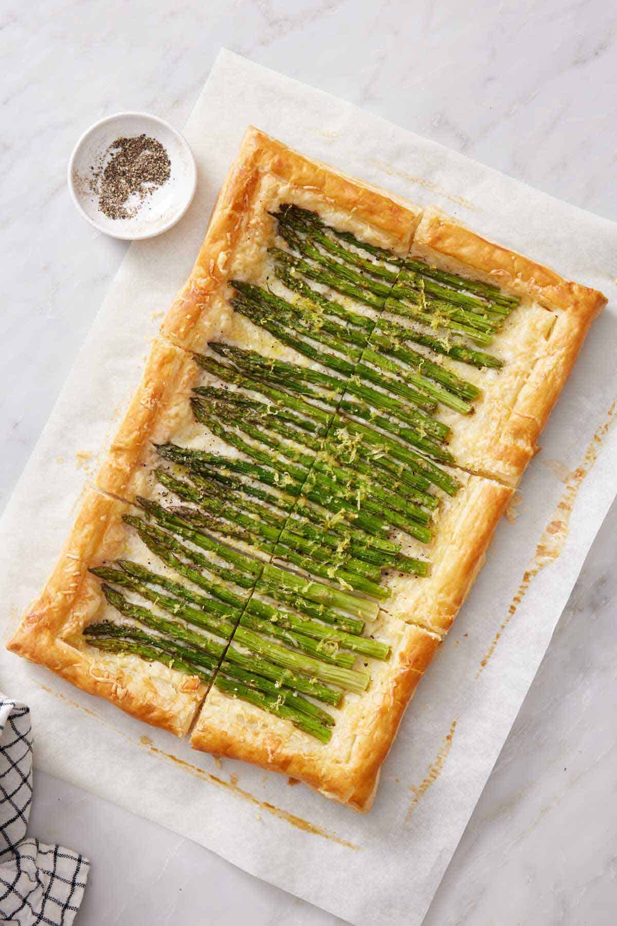 Overhead view of a cut asparagus tart on a sheet of parchment with a bowl of pepper on the side.