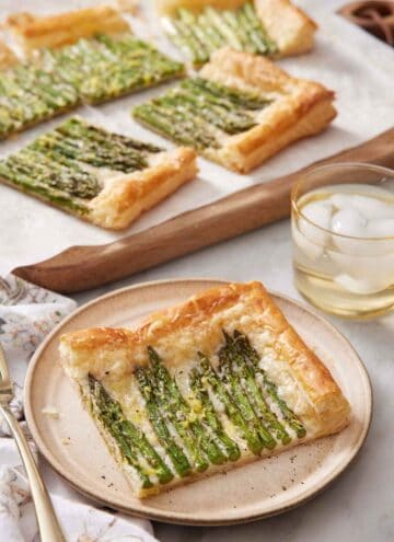 A plate with a slice of asparagus tart with the rest in the back on a serving board along with an iced drink.