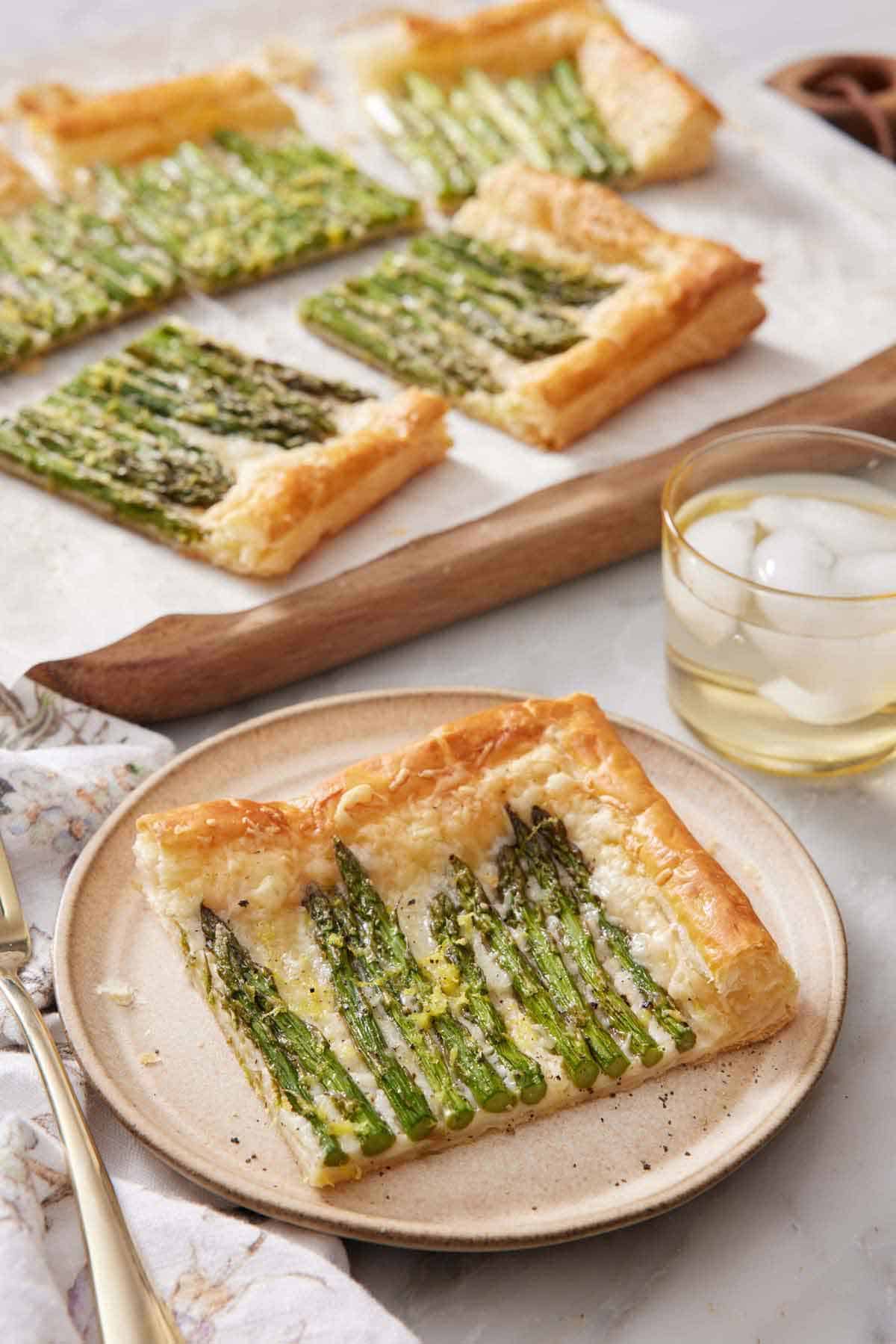 A plate with a slice of asparagus tart with the rest in the back on a serving board along with an iced drink.