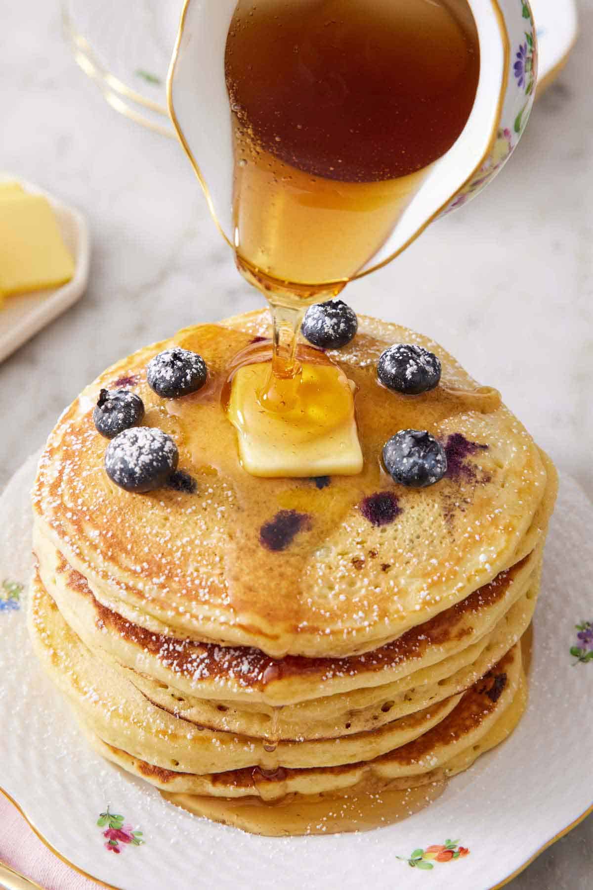A stack of blueberry pancakes topped with butter with maple syrup poured on top.