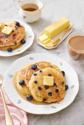 A plate with three blueberry pancakes topped with maple syrup, butter, and more blueberries. A coffee, butter, maple syrup, and another stack of blueberry pancakes in the background.