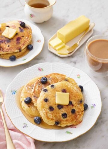 A plate with three blueberry pancakes topped with maple syrup, butter, and more blueberries. A coffee, butter, maple syrup, and another stack of blueberry pancakes in the background.