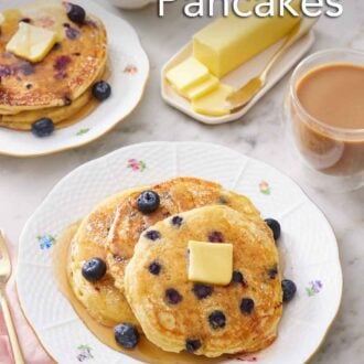 Pinterest graphic of a plate with three blueberry pancakes topped with maple syrup, butter, and more blueberries. A coffee, butter, maple syrup, and another stack of blueberry pancakes in the background.
